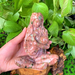 May include: A pink and green stone carving of a mother holding a baby. The carving is detailed and intricate, with the mother's hair flowing down her back and the baby nestled in her arms.