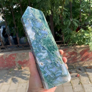 Puede incluir: Un obelisco de cristal rectangular con una superficie pulida. La piedra presenta una mezcla de patrones azul claro, blanco y verde. El obelisco se sostiene en una mano, mostrando su tamaño y forma.