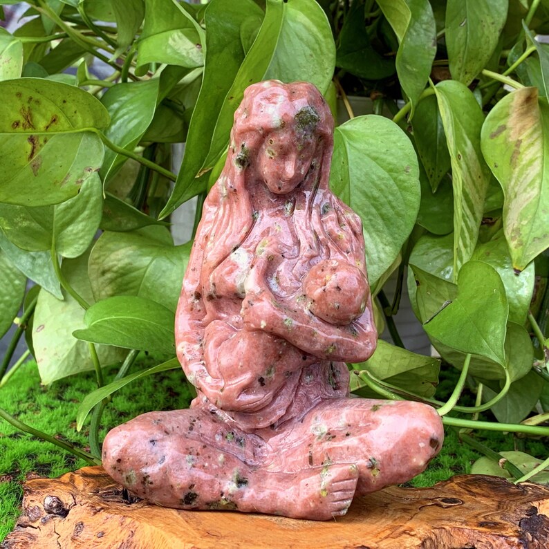 May include: A pink and green stone sculpture of a mother holding a baby. The sculpture is carved in a realistic style and has a natural, earthy look.