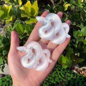 May include: Two white iridescent snake sculptures made of polished stone. The snakes are curled up in a coil shape.