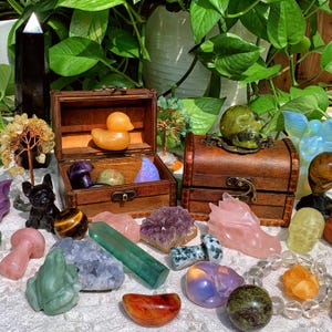 Pode incluir: Uma cole&ccedil;&atilde;o de esculturas de cristal e pedras preciosas coloridas, incluindo um pato, uma caveira, um drag&atilde;o e um cogumelo. As esculturas est&atilde;o dispostas sobre uma superf&iacute;cie branca com um ba&uacute; de madeira e plantas verdes no fundo.