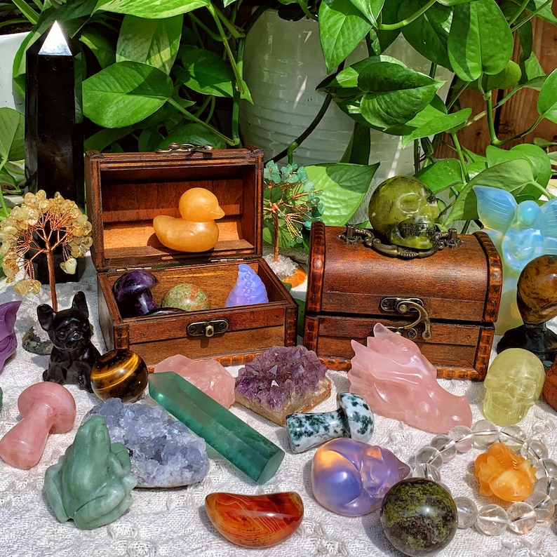 Pode incluir: Uma cole&ccedil;&atilde;o de esculturas de cristal e pedras preciosas coloridas, incluindo um pato, uma caveira, um drag&atilde;o e um cogumelo. As esculturas est&atilde;o dispostas sobre uma superf&iacute;cie branca com um ba&uacute; de madeira e plantas verdes no fundo.