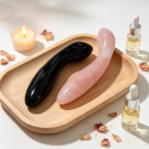 May include: Two smooth, curved massage wands, one crafted from polished black stone and the other from light pink rose quartz, resting on a rectangular wooden tray alongside a small lit candle and two glass dropper bottles of oil.