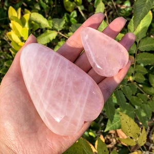 May include: Two pink rose quartz heart-shaped stones, one larger than the other, are held in a hand. The stones are smooth and polished.