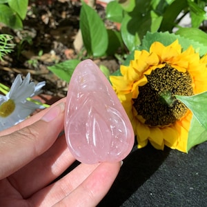 May include: A pink rose quartz carving of a goddess figure. The carving is smooth and polished, with intricate details. The carving is held in a hand against a background of green foliage and a yellow sunflower.