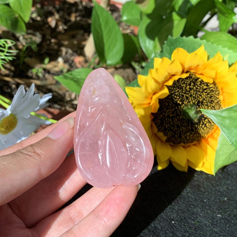 May include: A pink rose quartz carving of a goddess figure. The carving is smooth and polished, with intricate details. The carving is held in a hand against a background of green foliage and a yellow sunflower.