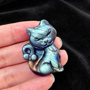 Puede incluir: Una piedra preciosa de labradorita pulida tallada con la forma de un gato sentado. La piedra muestra destellos iridiscentes azules, verdes y dorados en su superficie, con detalles grabados que definen la cara, las orejas y la cola del gato.