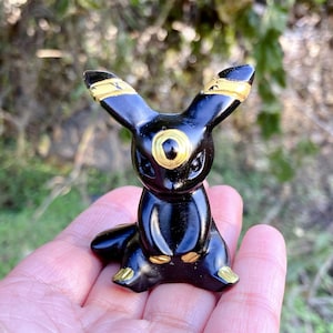 Könnte beinhalten: Glänzende schwarze Figur einer Kreatur mit großen Ohren und einem runden, goldenen Auge. Die Figur hat goldene Akzente an Ohren und Füßen. Die Figur sitzt, die Ohren zeigen nach oben. Die Figur wird in einer Hand gehalten.