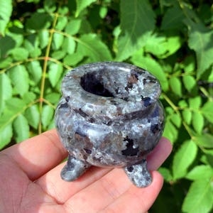 Puede incluir: Un pequeño caldero de piedra gris y negro moteado con tres patas. El caldero está hecho de piedra y tiene un acabado liso y pulido.