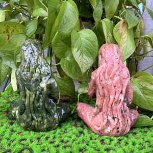 Puede incluir: Dos peque&ntilde;as esculturas de piedra de figuras sentadas de espaldas al espectador. La figura de la izquierda es verde con remolinos blancos, y la figura de la derecha es rosa con motas blancas.