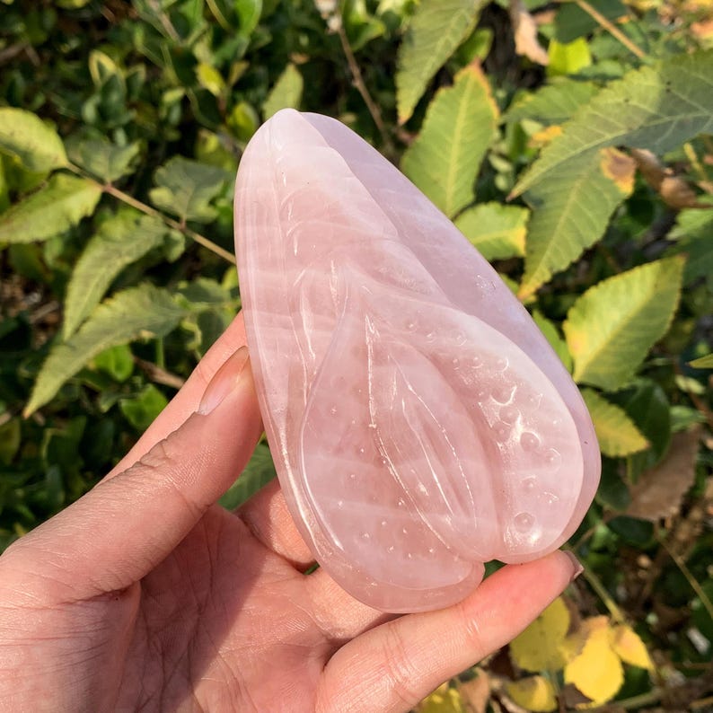 May include: A carved pink rose quartz crystal in the shape of a heart. The crystal is smooth and polished, with a delicate floral design.