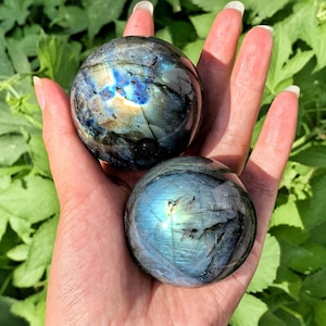 Può includere: Due sfere di cristallo di labradorite iridescenti, una blu scuro e una blu chiaro, con un bagliore dorato, sono tenute in una mano.