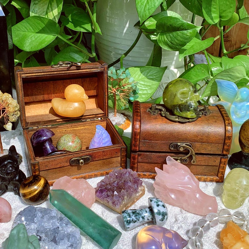 Pode incluir: Dois ba&uacute;s de madeira cheios de v&aacute;rios cristais, pedras e estatuetas. Os ba&uacute;s est&atilde;o abertos e cont&ecirc;m itens como uma estatueta de pato amarelo, um cogumelo roxo, uma pedra verde, um cristal roxo e uma caveira verde. Os ba&uacute;s s&atilde;o cercados por outros cristais e pedras, incluindo um grande aglomerado de ametista, uma estatueta de drag&atilde;o rosa e uma pedra olho de tigre.