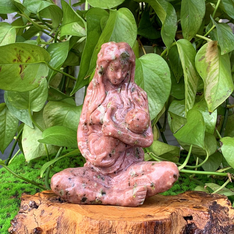 May include: A pink and green stone sculpture of a mother holding a baby. The sculpture is carved in a realistic style and has a smooth, polished finish.