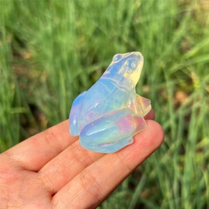Puede incluir: Una pequeña figura de rana de ópalo iridiscente. La rana está sentada sobre sus patas traseras con las patas delanteras dobladas. El ópalo es de color azul claro con un brillo arcoíris.