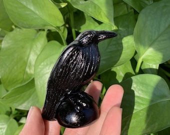 May include: Black obsidian crow figurine perched on a round base. The crow is carved with detailed feathers and has a realistic appearance.