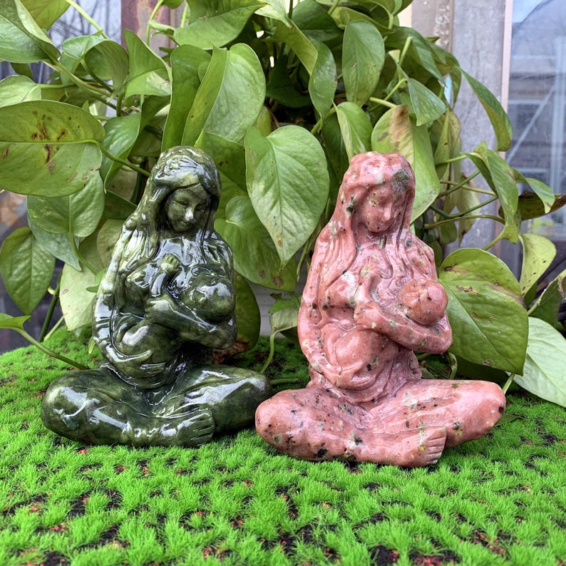 Puede incluir: Dos esculturas de piedra de una madre sosteniendo a un ni&ntilde;o. La escultura de la izquierda es verde y la escultura de la derecha es rosa.