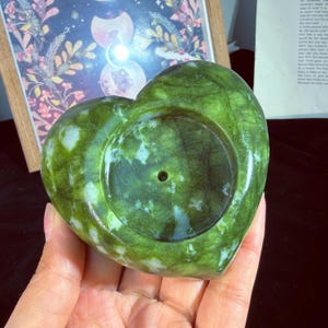 May include: A heart-shaped, green jade candle holder. The stone has a natural, mottled pattern with shades of green and white. The candle holder has a circular indentation to hold a candle. The item is held in a person's hand.