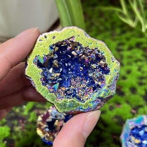 Op de afbeelding: Een geode met een levendig, iriserend interieur en een limoengroene en gouden buitenrand. Het interieur heeft een diepblauwe tint met sprankelende, veelkleurige kristalformaties. De geode wordt in een hand gehouden en toont zijn natuurlijke schoonheid.