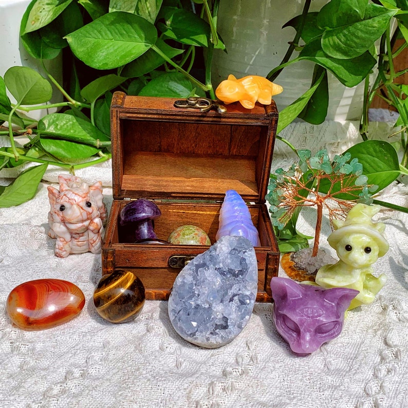 Pode incluir: Um ba&uacute; de madeira cheio de v&aacute;rios cristais e estatuetas. O ba&uacute; est&aacute; aberto e os itens dentro incluem um cogumelo roxo, um cristal azul, um cristal verde, uma estatueta amarela, uma estatueta branca e uma estatueta roxa.