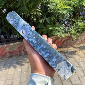 Puede incluir: Un obelisco rectangular de cristal de ágata musgosa. La piedra presenta patrones azules, blancos y verdes. El obelisco se sostiene en una mano, mostrando su superficie pulida y variaciones naturales. Mide unos 20 cm de largo.
