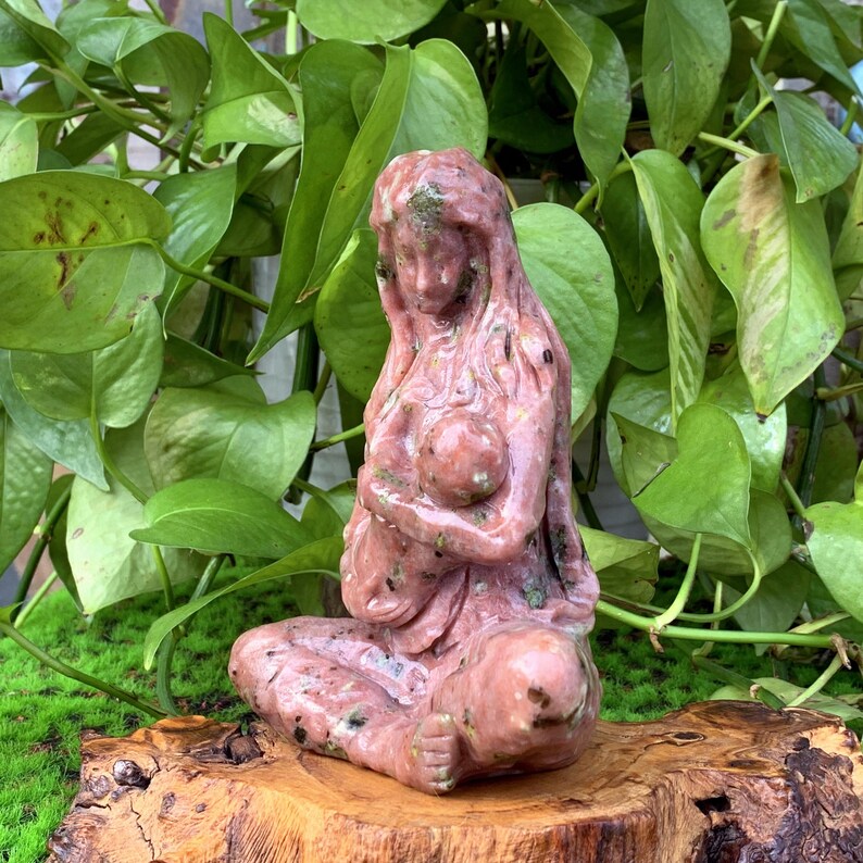 May include: A pink and green stone sculpture of a mother holding a child. The sculpture is carved in a realistic style and has a smooth, polished finish.