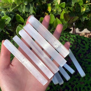 May include: A set of six white selenite wands, also known as selenite sticks, are arranged in a hand. The wands are smooth and have a natural, striated texture.