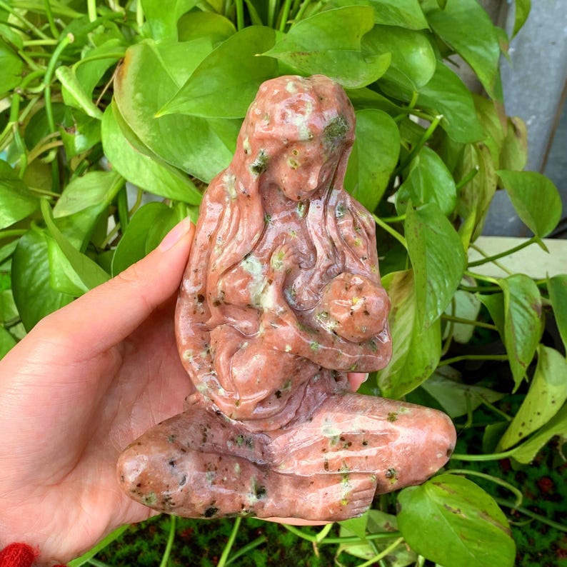 May include: A hand-carved stone sculpture of a mother and child. The sculpture is made of pink and green speckled stone and depicts a mother cradling her child in her arms.