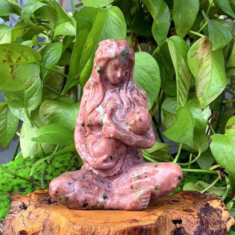 May include: A pink and green stone sculpture of a mother holding a baby. The sculpture is carved in a realistic style and has a smooth, polished finish.