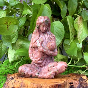 May include: A pink and green stone sculpture of a mother holding a baby. The sculpture is carved in a realistic style and has a smooth, polished finish.