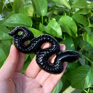 May include: Black obsidian snake sculpture. The snake is curled in a coil with its head raised. The sculpture is polished and has a shiny finish.