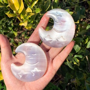 May include: Two white crescent moon shaped crystal carvings with a sleeping face and stars carved into the surface.