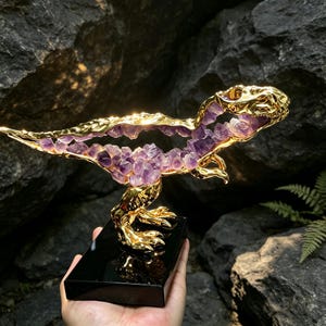 May include: A Tyrannosaurus rex sculpture featuring a polished gold-tone metal frame filled with clusters of purple amethyst crystals. The dinosaur figure is mounted on a rectangular black base, showcasing a detailed skeletal structure.