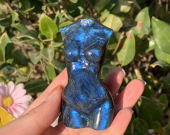 Pode incluir: Um cristal de labradorita azul esculpido na forma de um torso humano. O cristal tem um brilho iridescente e é polido liso.
