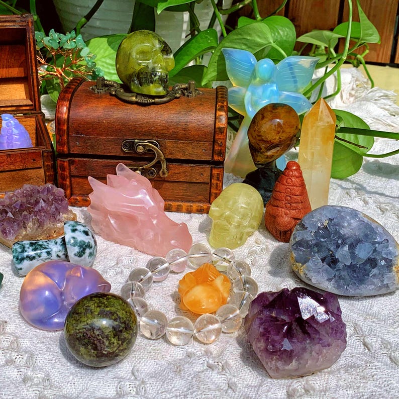 Pode incluir: Uma cole&ccedil;&atilde;o de v&aacute;rios cristais e pedras, incluindo um drag&atilde;o rosa, um cr&acirc;nio verde, um bracelete de quartzo transparente, uma ametista roxa, uma celestita azul e uma citrina amarela. Os cristais est&atilde;o dispostos sobre uma superf&iacute;cie branca com um ba&uacute; de madeira no fundo.