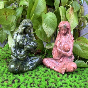 Puede incluir: Dos esculturas de piedra, una verde y otra rosa, que representan a madres sosteniendo beb&eacute;s. Las esculturas est&aacute;n talladas de forma realista y tienen un aspecto natural y terroso.