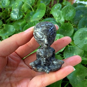 May include: A small, grey, carved stone figurine of an alien sitting in a meditative pose. The alien has large eyes and a detailed face.