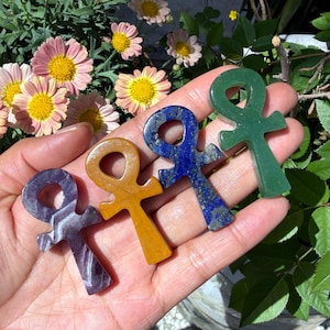 May include: Four ankh-shaped gemstone pendants in shades of purple, yellow, blue, and green. The pendants are held in a hand, with flowers and greenery in the background. The ankh is an ancient Egyptian symbol of life.