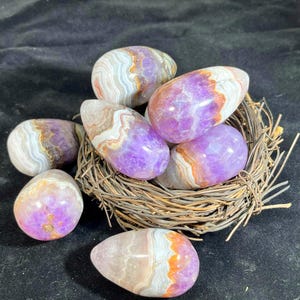 Op de afbeelding: Een verzameling gepolijste amethiststenen in verschillende vormen en maten, genesteld in een klein geweven nest. De stenen vertonen een reeks kleuren, waaronder paars, wit en oranje, met unieke bandpatronen. De achtergrond is een donker, getextureerd oppervlak.