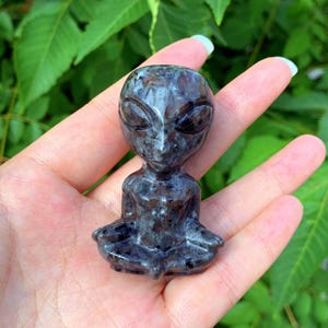 May include: A small, hand-carved alien figurine made of black and green speckled stone. The alien is sitting in a meditative pose with its hands resting on its lap.