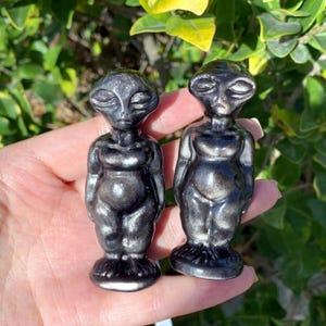 Op de afbeelding: Twee kleine, grijze, buitenaards gevormde beeldjes met een metallic glans. De beeldjes staan ​​recht met hun armen langs hun zijden. Ze hebben grote hoofden, grote ogen en kleine monden.