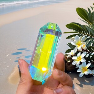 May include: A hand holds a faceted, iridescent crystal prism with a yellow core. The crystal reflects green and blue hues. The background shows a sandy beach, ocean, and white flowers. The crystal is approximately 10 cm long.