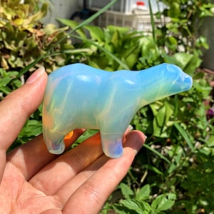 Pode incluir: Uma pequena figura de urso de opala esculpida à mão. O urso está de pé sobre suas quatro patas e tem uma cor iridescente azul claro e branco.