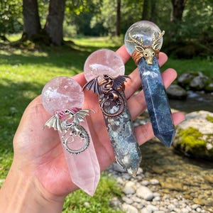 May include: Three crystal wands with spherical tops. The left is light pink with a silver dragon. The center is mottled green and gray with a copper dragon. The right is dark blue-green with a gold deer head. Each has a clear crystal sphere.
