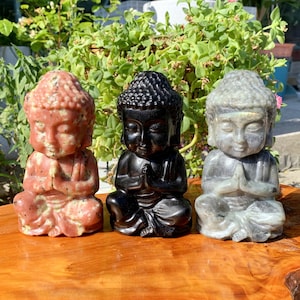 Peut inclure: Trois petites statues de Bouddha en méditation sculptées dans différents types de pierre. Le Bouddha de gauche est rose, le Bouddha du milieu est noir et le Bouddha de droite est gris.