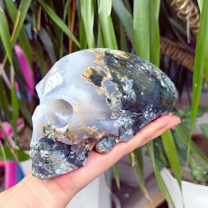 May include: A polished stone carving in the shape of a human skull, featuring translucent light gray agate with prominent dark green, black, and golden-brown moss-like inclusions throughout the surface, showcasing natural mineral patterns.