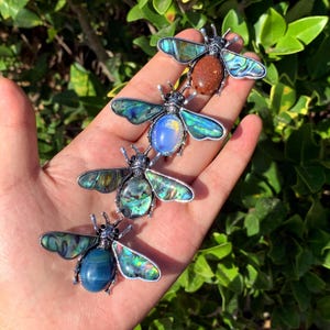 May include: Four insect brooches with iridescent wings and gemstone bodies. The brooches are silver-toned metal with bodies in shades of blue, brown, and iridescent green. The brooches are displayed against a background of green foliage.