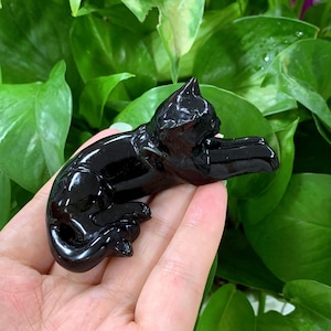 Könnte beinhalten: Schwarze Obsidian-Katzenfigur. Die Katze liegt auf der Seite mit dem Kopf zur Seite gedreht. Die Figur ist poliert und hat eine glänzende Oberfläche.