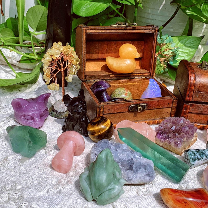 Pode incluir: Uma cole&ccedil;&atilde;o de cristais, pedras e estatuetas dispostas sobre uma superf&iacute;cie de tecido branco. Os itens incluem uma estatueta de gato roxo, uma estatueta de gato preto, uma estatueta de sapo verde, uma estatueta de cogumelo rosa, um cristal verde, um aglomerado de ametista roxo, uma pedra de olho de tigre, um pato de borracha amarelo e uma caixa de madeira com um cristal azul dentro.