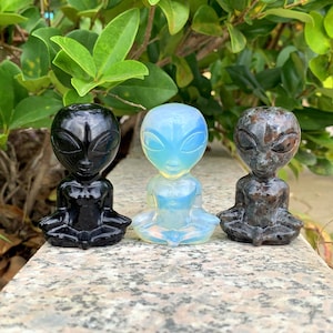 May include: Three alien figurines in a row, each in a seated pose. The first is black, the second is opalescent blue and white, and the third is a mottled grey. The figurines are on a stone surface with green foliage in the background.